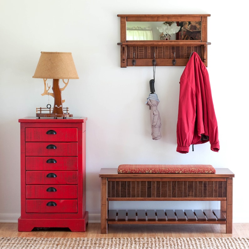 May include: A red wooden chest of drawers with six drawers and black handles. A wooden bench with a red cushion and a shelf underneath. A wooden wall mounted mirror with hooks and a red jacket hanging on one of the hooks. A lamp with a brown shade and a wooden base shaped like a tree.