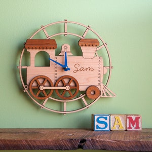 May include: Wooden train-shaped clock with blue hands and the name "Sam" written on the front. The clock is mounted on a wooden circle with a train track design. The clock is sitting on a wooden shelf with three wooden blocks that spell out "SAM".