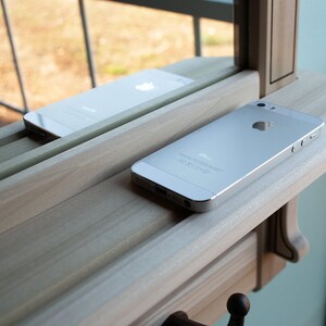 May include: Two silver iPhones on a wooden shelf. The top iPhone is reflected in a mirror behind it. The shelf has a decorative edge and a hook on the bottom.