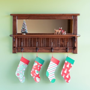 May include: A wooden shelf with a mirror and four hooks. There are four Christmas stockings hanging from the hooks. The stockings are red and green with different patterns. There is a small Christmas tree on the shelf.