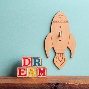 May include: A wooden rocket-shaped light switch cover with a star and circle design, mounted on a light blue wall. The word "DREAM" is spelled out with colorful wooden blocks on a wooden shelf.