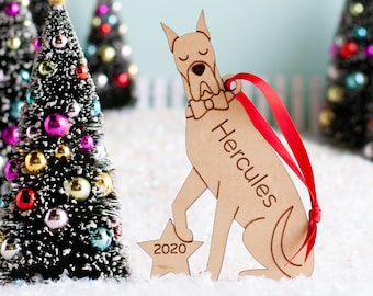May include: A wooden Christmas ornament shaped like a Great Dane dog holding a star with the year 2020 on it. The ornament is personalized with the name "Hercules" and has a red ribbon attached.