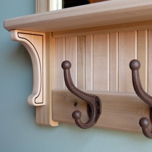 May include: A wooden coat rack with a shelf and two brown metal hooks. The rack has a decorative trim and is made of light wood.