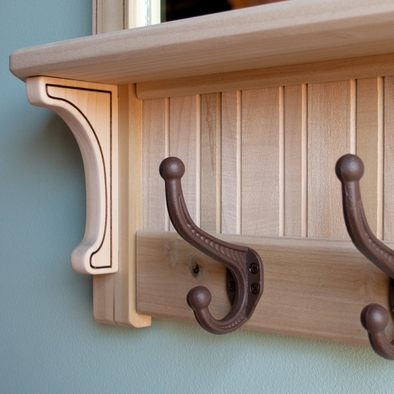 May include: A wooden coat rack with a shelf and two brown metal hooks. The rack has a decorative trim and is made of light wood.