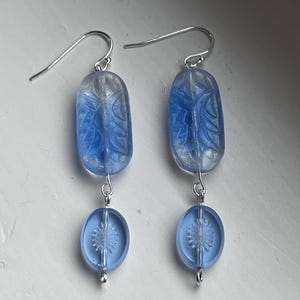May include: A pair of blue and clear glass earrings. Each earring features a long, oval-shaped bead with a leaf design, connected to a smaller, oval bead with a sunburst pattern. The earrings have silver-colored ear wires.
