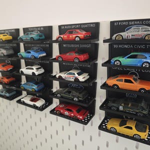Könnte beinhalten: Eine Sammlung von Miniatur-Spielzeugautos, die auf schwarzen Regalen an einer weißen Wand ausgestellt sind. Die Autos sind in verschiedenen Farben, darunter Gelb, Rot, Blau und Weiß. Jedes Regal ist mit Marke und Modell des Autos beschriftet.