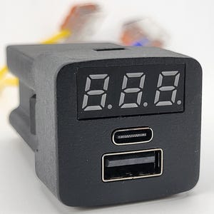 May include: A black rectangular electronic module featuring a digital LED display screen at the top, a USB-C port in the center, and a standard USB-A port at the bottom. The device has a matte finish and includes attached yellow and orange wiring.