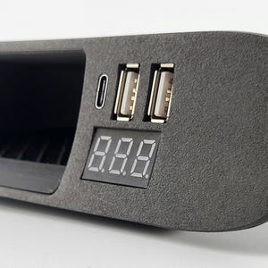 Puede incluir: Un panel de consola electrónica de color gris oscuro texturizado que cuenta con un puerto USB-C, dos puertos USB-A y una pantalla digital rectangular. El dispositivo tiene un acabado mate y un compartimento de almacenamiento empotrado.