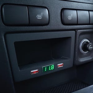 Op de afbeelding: Close-up van een auto dashboard met een ingebouwde USB-oplaadpoort en een digitaal display met "11.8". Het dashboard heeft ook knoppen voor stoelverwarming en een sigarettenaansteker.