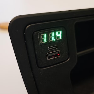 Puede incluir: Dispositivo electrónico negro con una pantalla digital verde que muestra el número 8.8.9. Debajo de la pantalla hay dos puertos, uno USB y otro USB-C. El dispositivo está integrado en una carcasa de plástico negro.