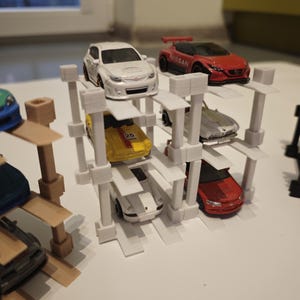 Könnte beinhalten: Eine Sammlung von Spielzeugautos in verschiedenen Farben, darunter Weiß, Rot, Gelb und Silber, die auf einem weißen, mehrstöckigen Aufbewahrungsregal ausgestellt sind. Die Autos sind Miniaturrepliken verschiedener Modelle.