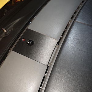 May include: A dark gray automotive dashboard panel featuring a central black square insert. This insert contains a small, circular black button and a single, illuminated red LED indicator light. The panel has a smooth, matte finish and integrated vents.