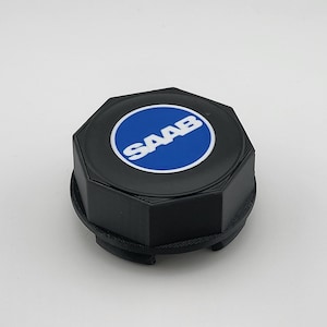 May include: A black octagonal wheel centre cap featuring a circular blue emblem in the centre with the white text SAAB printed across it. The cap has a textured side profile and a multi-pronged attachment mechanism on the underside for secure mounting.