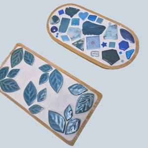 May include: Two decorative trays with wooden frames. One is rectangular with blue leaf designs. The other is oval, featuring a mosaic of blue and white tiles, including hearts and geometric shapes.