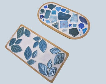 May include: Two decorative trays with wooden frames. One is rectangular with blue leaf designs. The other is oval, featuring a mosaic of blue and white tiles, including hearts and geometric shapes.