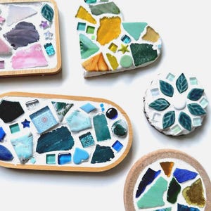 May include: A collection of handmade mosaic coasters and decorative pieces in various shapes. The pieces feature colorful glass shards in shades of blue, green, yellow, and pink, set in a white or tan base with wooden or tan borders.