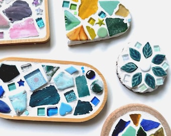 May include: A collection of handmade mosaic coasters and decorative pieces in various shapes. The pieces feature colorful glass shards in shades of blue, green, yellow, and pink, set in a white or tan base with wooden or tan borders.