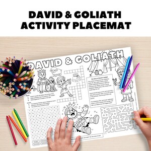 Op de afbeelding: Een witte activiteit placemat met de tekst "David & Goliath" en illustraties van het bijbelse verhaal. Inclusief een kruiswoordraadsel, doolhof en kleurgebieden. Gekleurde potloden en kleurpotloden zijn zichtbaar.