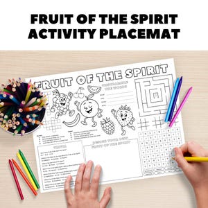 Op de afbeelding: Een witte activiteit placemat met de tekst "FRUIT OF THE SPIRIT ACTIVITY PLACEMAT". De placemat bevat fruitillustraties, een kruiswoordpuzzel, een doolhof en een woordzoeker. Gekleurde potloden en kleurpotloden zijn zichtbaar.