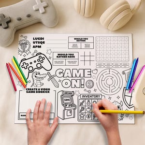 Pode incluir: Um tapete de atividades branco com tema de videogame, com um controle, um labirinto e as palavras "GAME ON!" e "CREATE A VIDEO GAME SIDEKICK". Lápis de cor e uma mão segurando um lápis amarelo são visíveis. Um controle de jogo cinza e fones de ouvido estão no fundo.
