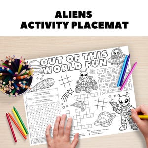 May include: A white activity placemat with the text "ALIENS ACTIVITY PLACEMAT" and "OUT OF THIS WORLD FUN". The placemat features alien-themed illustrations, a maze, a crossword puzzle, and word search. Colored pencils and crayons are visible.