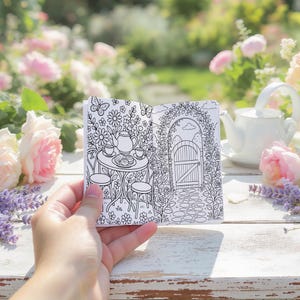 May include: A small, open coloring book held in a hand, featuring a black and white illustration of a garden tea party scene. The scene includes a table, chairs, a teapot, and a gate. The background is blurred with flowers and a white teapot.