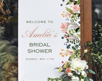 May include: A white sign on an easel with the text "WELCOME TO Amelia's BRIDAL SHOWER SUNDAY, MAY 11TH". The sign is decorated with watercolor floral elements in pink, green, and yellow. A bouquet of white flowers and greenery is at the base.