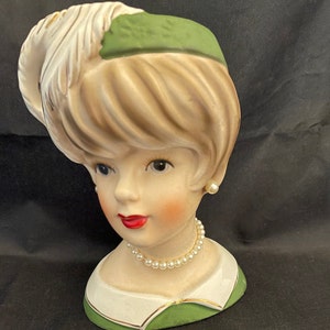 May include: A vintage ceramic bust of a woman with blonde hair, wearing a green hat with a white feather and a pearl necklace. The bust is a vase.
