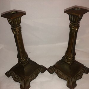 May include: A pair of antique brass candle holders with ornate details. The holders have a square base and a tall, slender column with a decorative top.