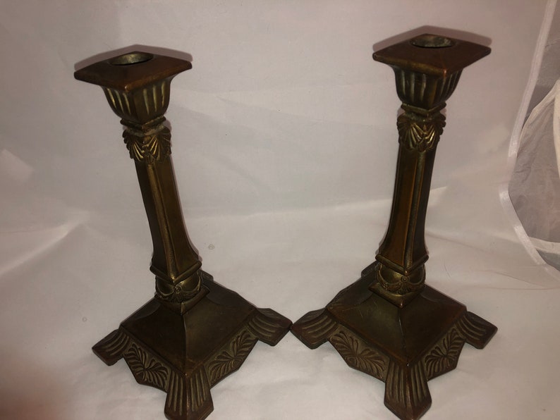May include: A pair of antique brass candle holders with ornate details. The holders have a square base and a tall, slender column with a decorative top.