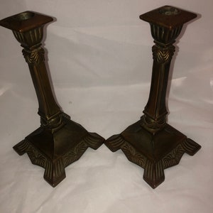 May include: A pair of antique bronze candle holders with ornate details. The holders have a square base with a decorative design and a tall, slender column with a square top.