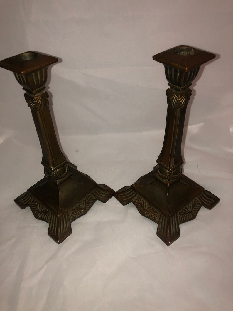 May include: A pair of antique bronze candle holders with ornate details. The holders have a square base with a decorative design and a tall, slender column with a square top.