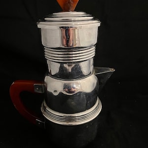 May include: A silver and brown Art Deco style coffee pot with a rounded body and a lid with a brown plastic knob. The pot has a handle made of brown plastic.
