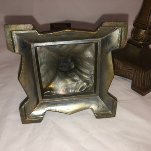 May include: A pair of antique brass candle holders with ornate details. The base of each holder is square with a decorative design. The candle holders are in good condition with some signs of age.