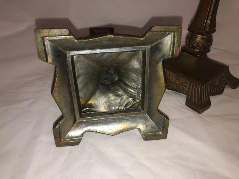 May include: A pair of antique brass candle holders with ornate details. The base of each holder is square with a decorative design. The candle holders are in good condition with some signs of age.