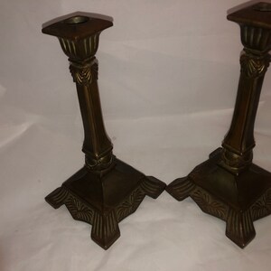 May include: A pair of antique brass candle holders with ornate details. The holders have a square base with a fluted design and a tall, slender column.