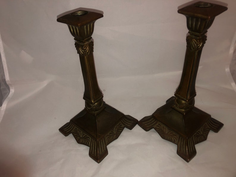 May include: A pair of antique brass candle holders with ornate details. The holders have a square base with a fluted design and a tall, slender column.