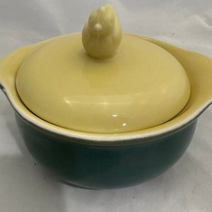 May include: A yellow ceramic casserole dish with a lid featuring a small bird finial. The dish is green on the inside and has a white rim.