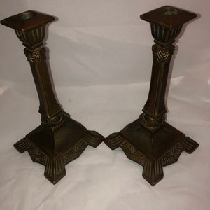 May include: A pair of antique brass candle holders with ornate details. The holders have a square base with a decorative design and a tall, slender stem.