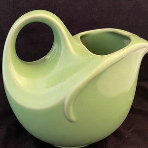 May include: A light green ceramic pitcher with a large, circular handle and a curved spout. The pitcher has a smooth, glossy finish and a rounded body. The design is simple and elegant, suitable for serving beverages.