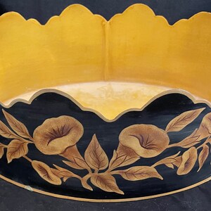 May include: A decorative oval-shaped serving tray with a scalloped edge. The tray features a black exterior with floral and leaf designs in shades of brown and gold. The interior is gold, and it has two gold handles.