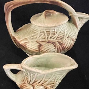 May include: A green and brown ceramic teapot and creamer set with a pine cone design. The teapot has a lid and a curved handle. The creamer has a handle and a rounded shape.