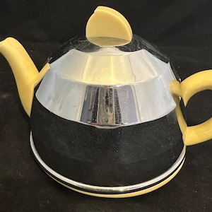 May include: A chrome and yellow teapot with a rounded, geometric design. The teapot has a lid with a rounded top and a handle on the side.