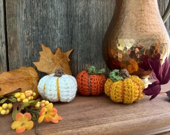 May include: Three crocheted pumpkins in white, orange, and yellow with green and brown accents sit on a wooden surface. Autumn leaves and a copper pitcher are in the background. The pumpkins are a fall-themed decoration.