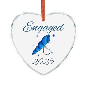Peut inclure: Ornement en forme de cœur transparent avec le mot "Engaged" en bleu. Un motif de coquillage bleu avec une bague et l'année 2025 est présenté en dessous. L'ornement est suspendu par un ruban orange.