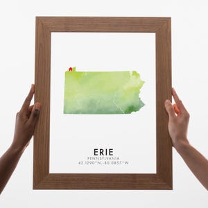 Könnte beinhalten: Ein gerahmter Druck mit einer Aquarellkarte von Pennsylvania in Grüntönen. Die Stadt Erie ist mit einem roten Haus-Symbol hervorgehoben. Der Text "ERIE PENNSYLVANIA 42.1290°N, -80.0857°W" ist darunter. Der Rahmen ist aus Holz.