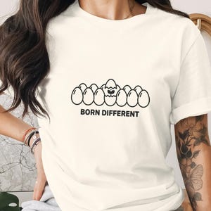 Op de afbeelding: Crèmekleurig T-shirt met een zwarte afbeelding van eieren, waarvan er één uitkomt. De tekst "BORN DIFFERENT" staat eronder. Het shirt heeft een casual stijl.