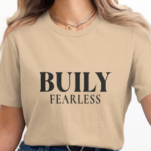 Puede incluir: Camiseta beige con las palabras "BUILY FEARLESS" impresas en negro. La camiseta tiene mangas cortas y cuello redondo. La modelo lleva vaqueros azules y un collar dorado.