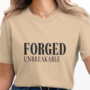 Puede incluir: Camiseta beige con la frase negra "FORGED UNBREAKABLE". La camiseta tiene mangas cortas y cuello redondo. El texto está en negrita, con una fuente sans-serif.