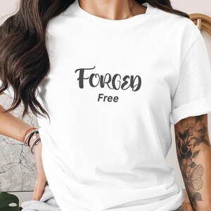 Peut inclure: T-shirt blanc avec l'inscription "Forged Free" en noir, dans une police manuscrite. Le t-shirt a des manches courtes et une coupe décontractée. L'image montre le t-shirt sur une personne.